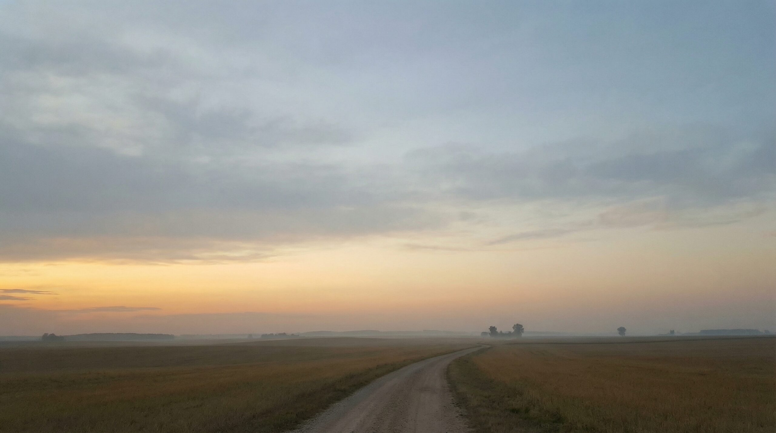 A straight dirt road extending toward a warm amber sunrise horizon across a wide flat plain under a soft blue-grey morning sky