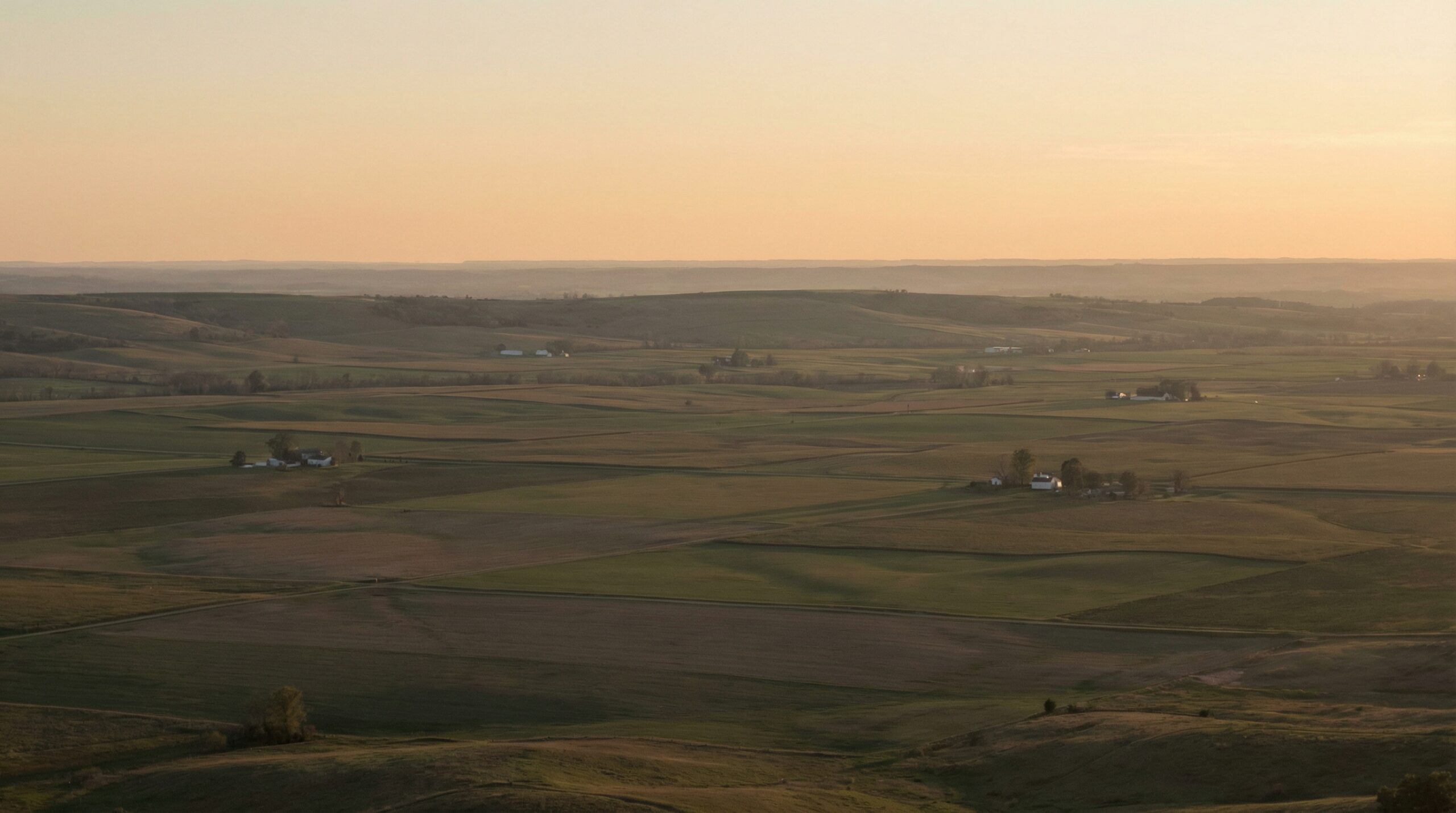 An aerial view of rolling farmland and patchwork fields stretching to the horizon in warm late-afternoon golden light