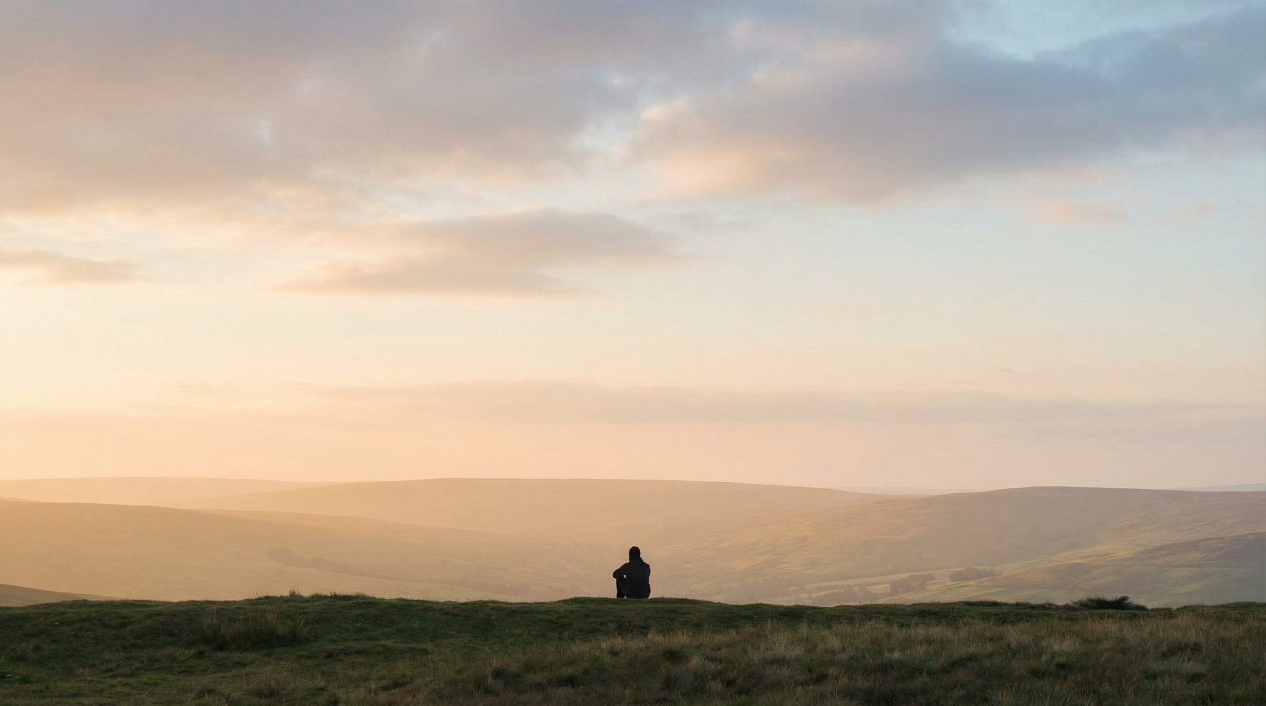 A lone figure seated on an open hillside in silhouette, facing a wide misty valley in soft golden evening light.
