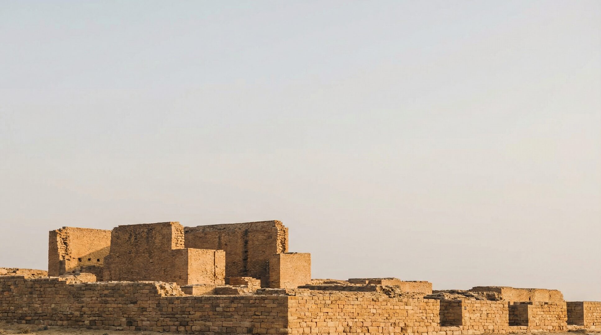 Ancient Mesopotamian brick ruins under a wide pale sky in warm afternoon light, weathered and incomplete against the open horizon