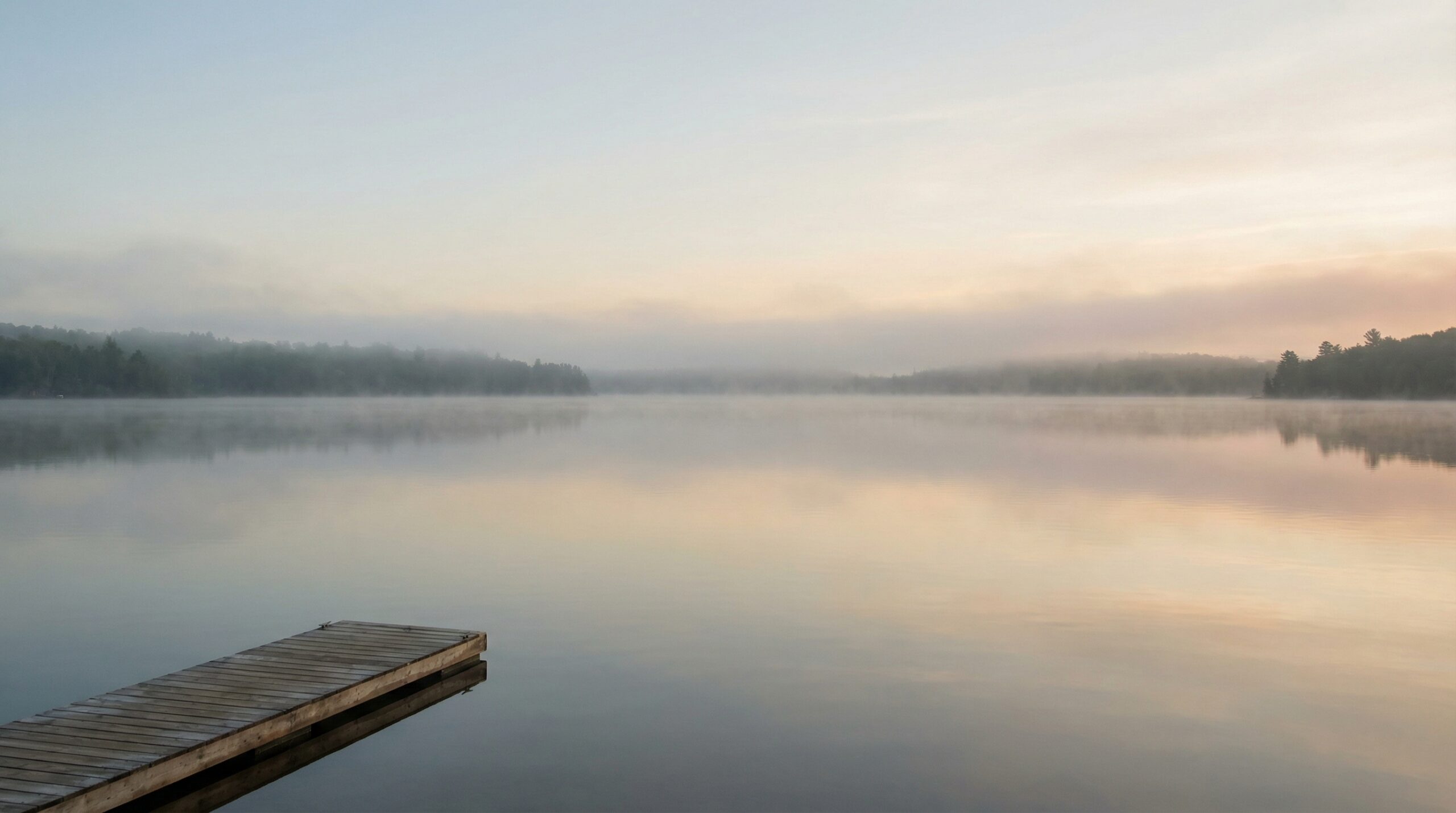 A small wooden dock extending into a still lake at early morning, with soft mist over the water and a warm peach and blue sky reflected in the surface