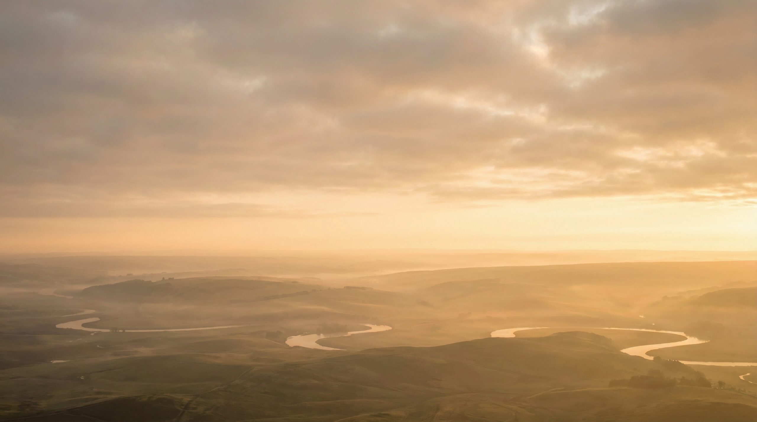 Warm amber dawn light spreading across a broad misty valley with a winding river below
